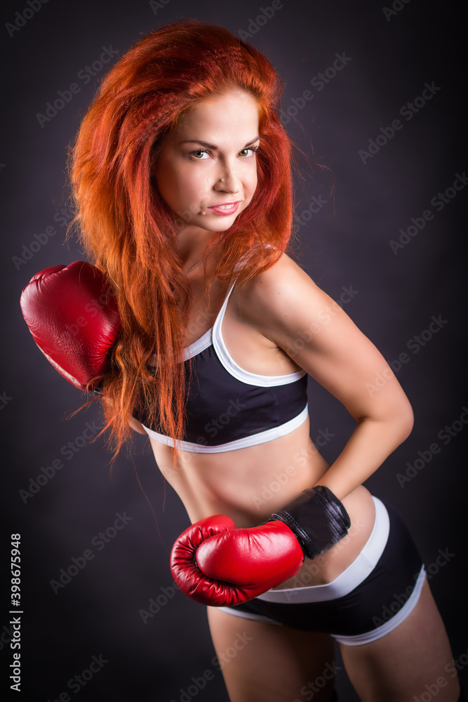 A beautiful slender and sexy girl with red hair in a sports top and short shorts in a Boxing glove. Photo in the Studio on a black background. The concept of beauty and sport.