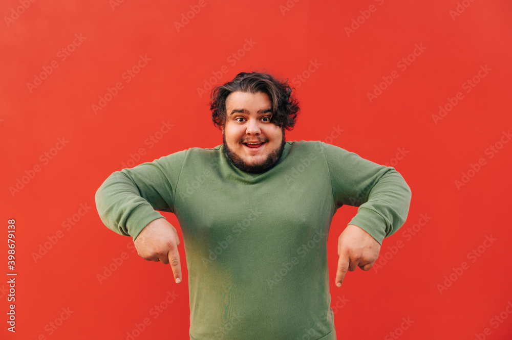 Cheerful and funny young bearded fat guy is standing on a red background and pointing his fingers down, indicating on a copy space, advertising. Cheerful and funny young bearded fat guy is standing on a red background and pointing his fingers down, indicating on a copy space, advertising.