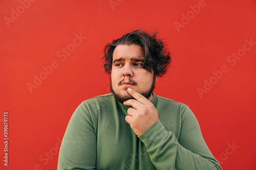 Closeup portrait of a thoughtful bearded hispanic man standing on a red background, expressing a thinking gesture, touching his chin with his hand.