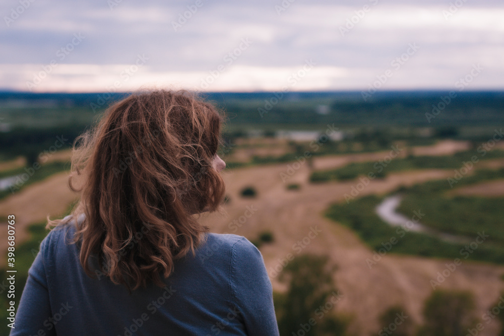 the girl sits on a hill and has a beautiful view of the river and fields in front of her