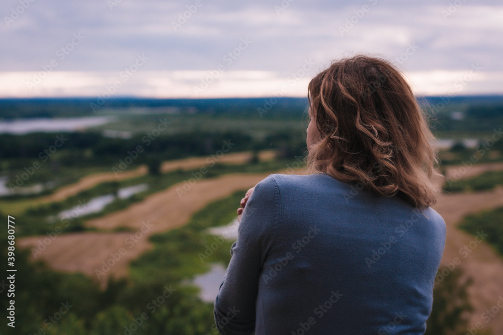 the girl sits on a hill and has a beautiful view of the river and fields in front of her