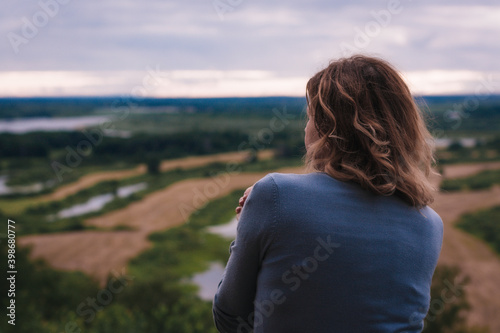 the girl sits on a hill and has a beautiful view of the river and fields in front of her