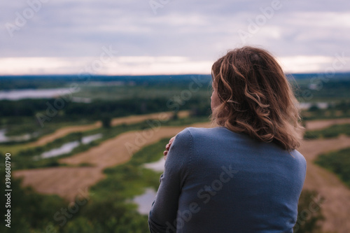 the girl sits on a hill and has a beautiful view of the river and fields in front of her