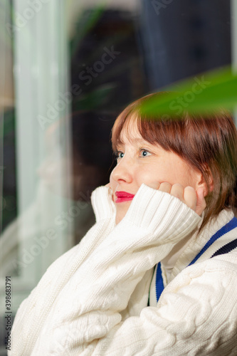 young pretty girl in a white sweater sitting on the windowsill at home by the window dreaming