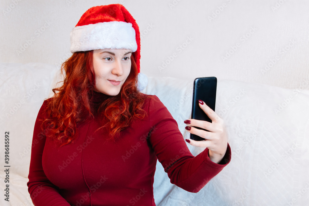 Red-haired young woman in Santa hat uses video chat on her mobile phone. Online greeting with Christmas. Red-haired young woman in Santa hat uses video chat on her mobile phone. Online greeting with Christmas.