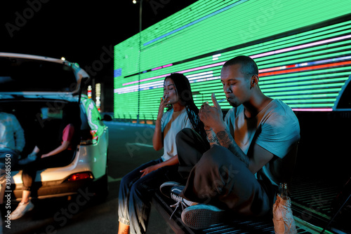 A group of two young casually dressed dark-skinned friends of different genders sitting in an opened car trunk lighting their cigarettes with a led screen and cars behind