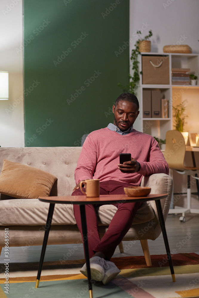African young man sitting on sofa in front of the table with cup of coffee and using his mobile phone during coffee break