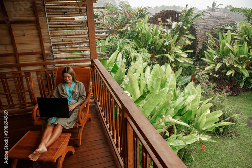 Freelance girl wering bathrobe sitting on the balcony working, typing on laptop during raining season on Bali island