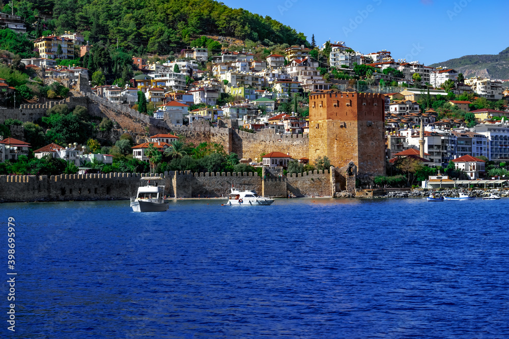 Two white boats near the medieval stone wall and the red Kizil Kule tower in Alanya (Turkey). Panorama of the tourist town with blue water, green forest and modern buildings on the hillside