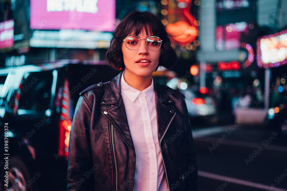 Half length of charming hipster girl dressed in leather jacket and stylish spectacles posing at urban setting in megalopolis, youthful female in trendy streetwear looking at camera on leisure Half length of charming hipster girl dressed in leather jacket and stylish spectacles posing at urban setting in megalopolis, youthful female in trendy streetwear looking at camera on leisure