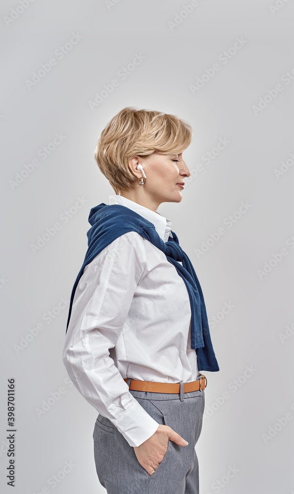 Elegant middle aged caucasian woman wearing business attire, smiling aside while using her earbuds, standing isolated over grey background