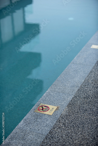No diving sign on a pool side for a shallow pool with a reflection showing a modern building; hotel or condominium, in a peaceful turquoise color scheme during the morning time.