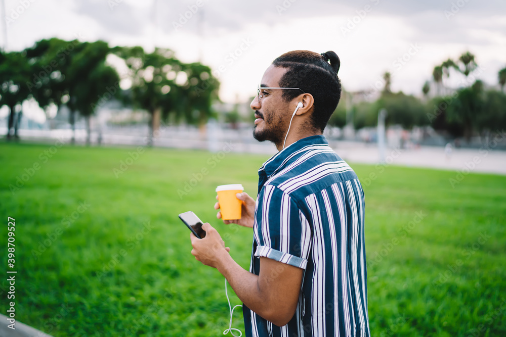 Fototapeta premium Rear view of positive dark skinned male holding smartphone listening music and walking with coffee to go cup,smiling african american hipster guy sending messages strolling in earphones enjoy playlist