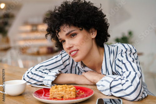 Beautiful happy curly woman smiling while drinking coffee with cake