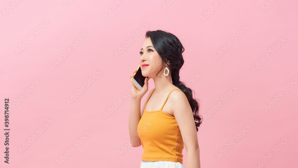 Portrait of a playful beautiful woman in summer clothes talking on mobile phone while standing and looking away isolated over pink background Portrait of a playful beautiful woman in summer clothes talking on mobile phone while standing and looking away isolated over pink background