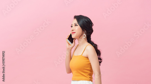 Portrait of a playful beautiful woman in summer clothes talking on mobile phone while standing and looking away isolated over pink background