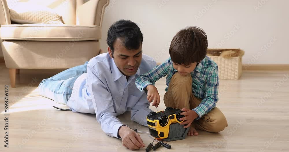 In warm living room Indian father his little son repairing toy car play together auto mechanic, child use screwdriver fixing broken truck with dad. Skills transmission, share knowledge, hobby concept