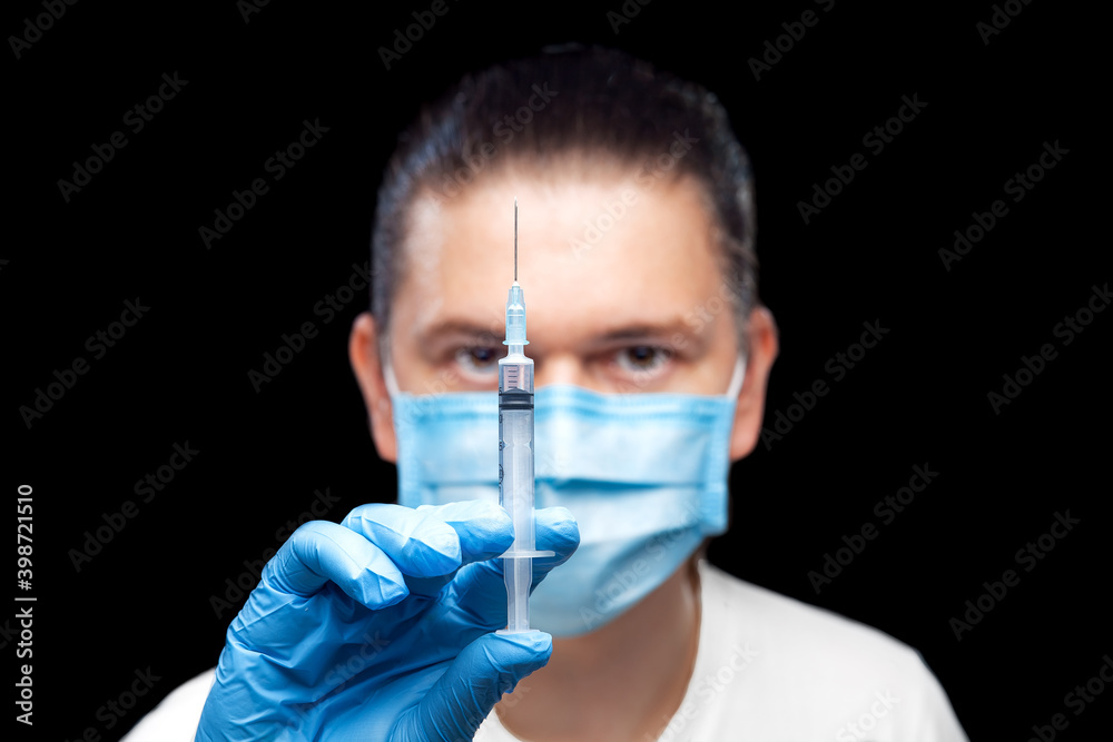 doctor in protective mask and sterile gloves holds syringe with vaccine for injection against virus in his hands and looks carefully assesses medical product close up isolated on black background. doctor in protective mask and sterile gloves holds syringe with vaccine for injection against virus in his hands and looks carefully assesses medical product close up isolated on black background.