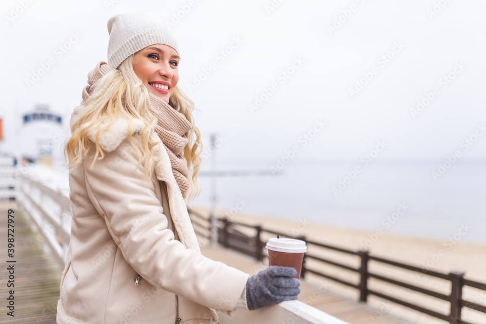 Cute middle-aged blonde woman enjoy time outdoor at winter day. Female wearing a light jacket, hat, scarf and have hot beverage.