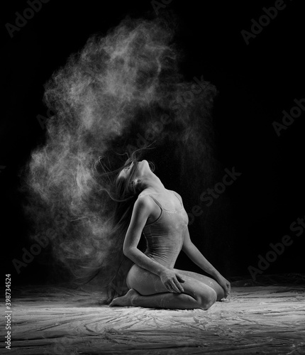 A beautiful slender ballet dancer girl wearing a bodysuit, sensually waves her long hair while sitting on the floor among the clouds of flying flour, on black. Artistic, commercial, monochrome design