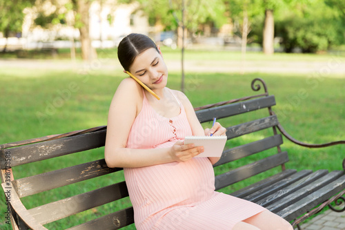 pregnant woman sits on a park bench and writes a to do list to the hospital and talking on the phone