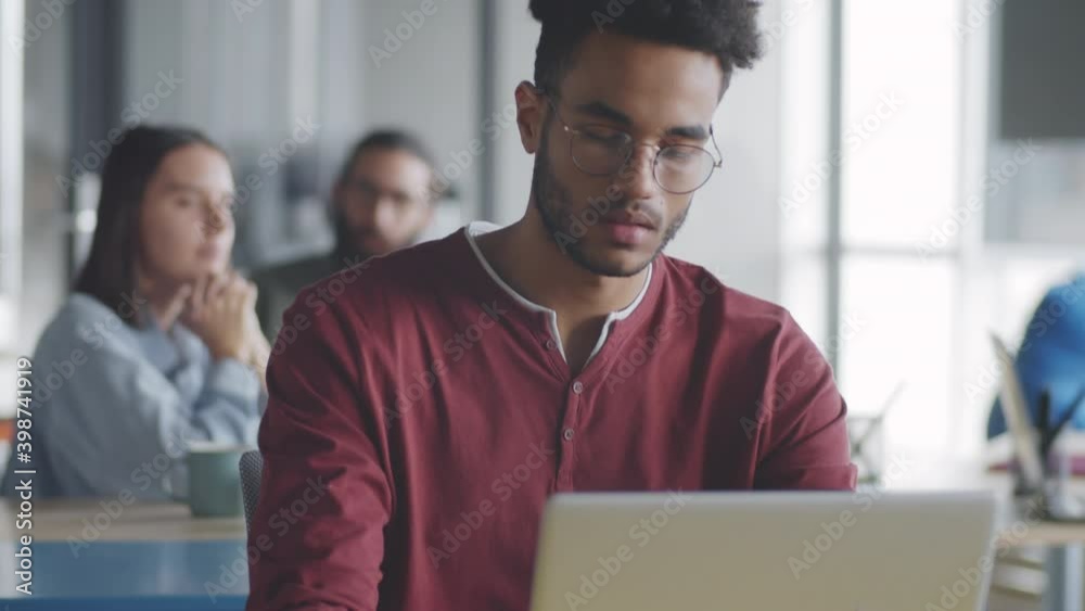 Tilt up portrait shot of young handsome mixed-race businessman in casualwear and glasses typing on laptop and then posing for camera while working at office desk Tilt up portrait shot of young handsome mixed-race businessman in casualwear and glasses typing on laptop and then posing for camera while working at office desk