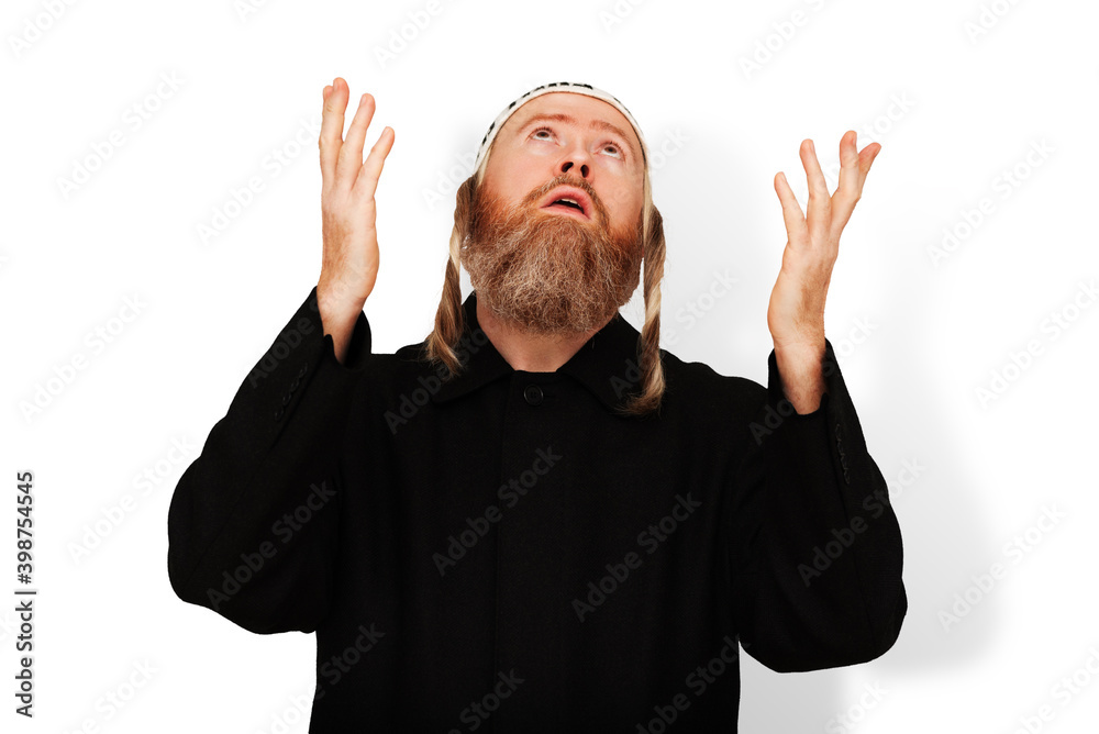 Praying bearded Jewish man with sidelocks in white kippah raising his hands to heaven. Studio portrait of hasid wearing black coat isolated on white background. Closeup.