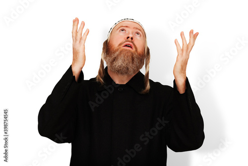 Obraz na płótnie Praying bearded Jewish man with sidelocks in white kippah raising his hands to heaven. Studio portrait of hasid wearing black coat isolated on white background. Closeup. w salonie