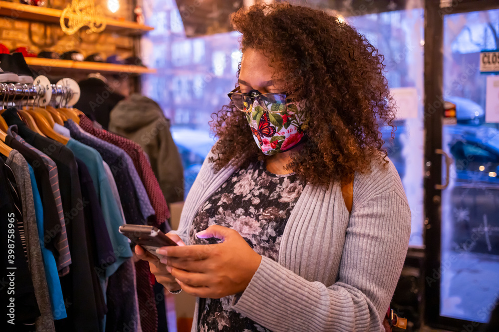 African American mixed woman with curly hair and wearing a protective mask is texting on her smart phone while shopping during Covid-19 pandemic. 