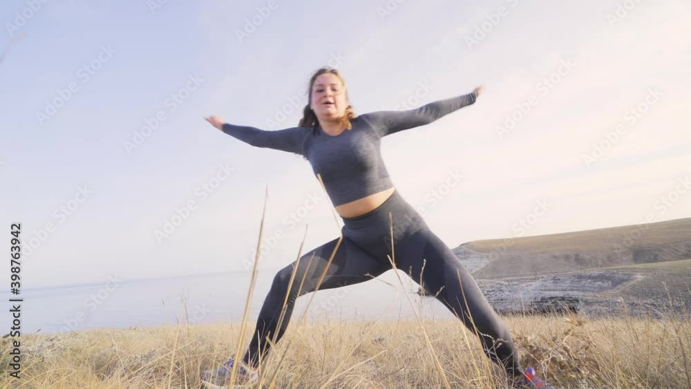 Plump woman with curly hair in tracksuit does exercise sitting on knee among sea coast dry grass at bright sunlight in early morning low angle shot Plump woman with curly hair in tracksuit does exercise sitting on knee among sea coast dry grass at bright sunlight in early morning low angle shot