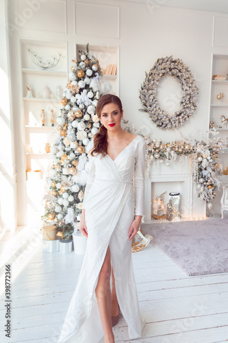 A girl stands against the background of a fireplace, decorated with a garland and a Christmas wreath, a girl in a white shiny dress. Pretty girl for christmas. Girl in a white dress and red lips.