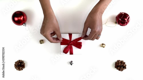 Hand and arm shot of a mixed race African American woman untying a bow on a small white gift box Christmas present wrapped with a red ribbon and bow sitting in middle of festive background.