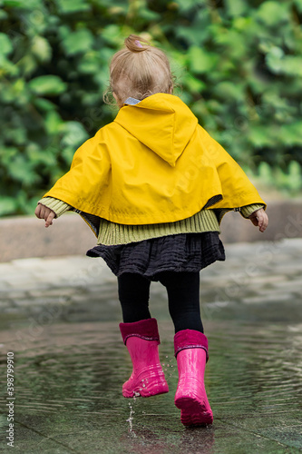 A pretty little girl in a yellow raincoat and pink rubber boots jumps on puddles with splashes and rejoices. Park, nature, outdoors. Universal Children's Day.
