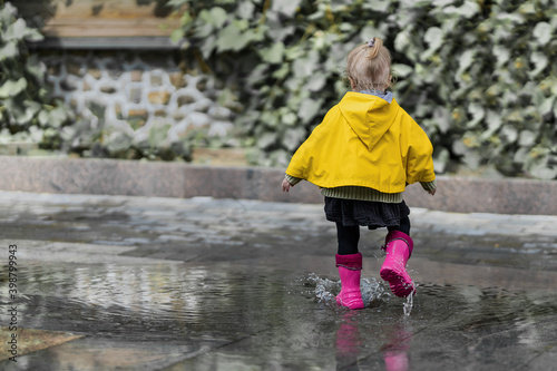 A nice little girl in a yellow raincoat and pink rubber boots jumps on puddles with splashes and rejoices. Park, nature, outdoors. Summer time. Universal Children's Day. childhood