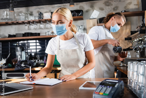 Waiter in medical mask writing in notebook near laptop, and barista working on background