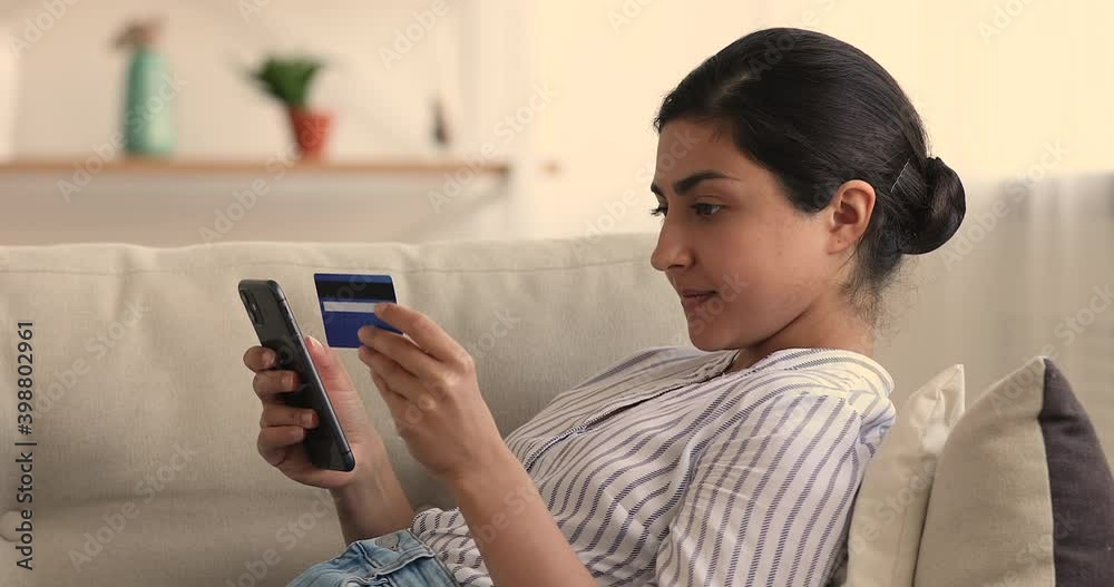 Side view satisfied Indian woman rest on sofa at home holds device enters credit card information use e-bank app on smartphone purchasing goods, services online. Virtual distant money transfer concept Side view satisfied Indian woman rest on sofa at home holds device enters credit card information use e-bank app on smartphone purchasing goods, services online. Virtual distant money transfer concept
