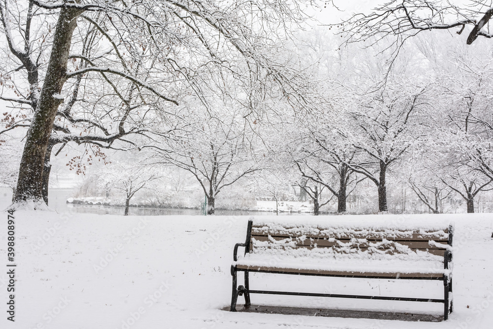 Wintry snow scene in a park with a snow-covered bench and a lake in the background.