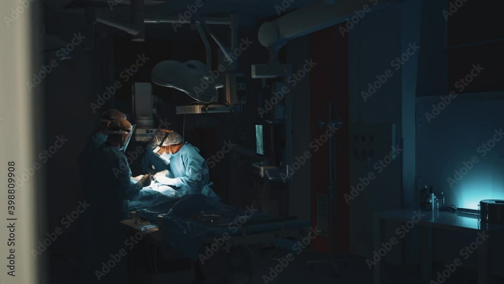View from the hall of the professional doctors wearing blue medical clothes in the dark operation theatre under the bright light of a surgical lamp doing operation for patient at the serious condition View from the hall of the professional doctors wearing blue medical clothes in the dark operation theatre under the bright light of a surgical lamp doing operation for patient at the serious condition