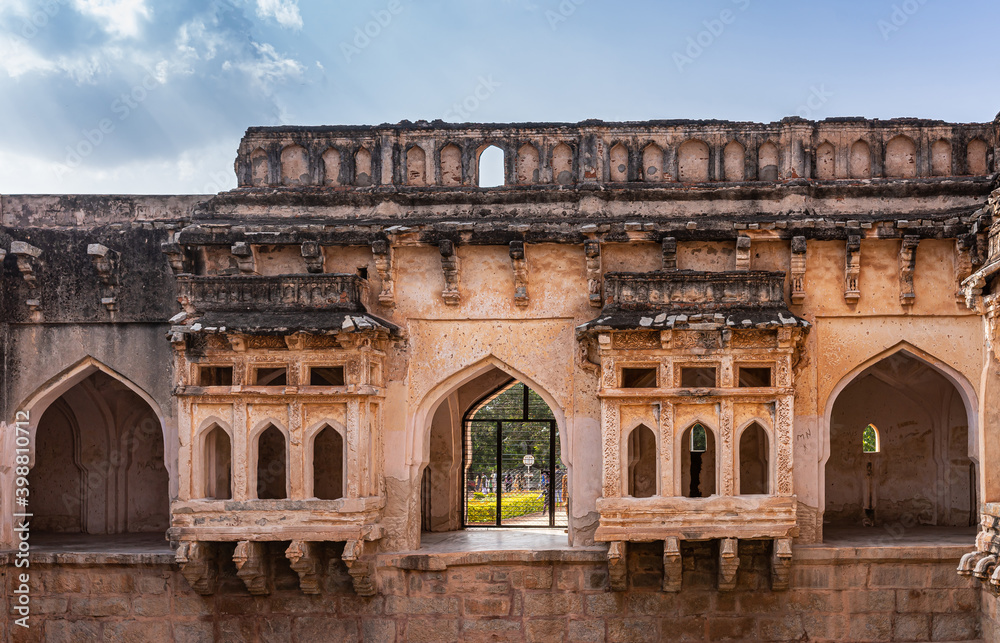 Hampi, Karnataka, India - November 4, 2013: Queens bath structure of Vijayanagara Empire. Closeup of 2 symetrical brown stone balconies portruding over pool. Corridor under blue cloudscape.