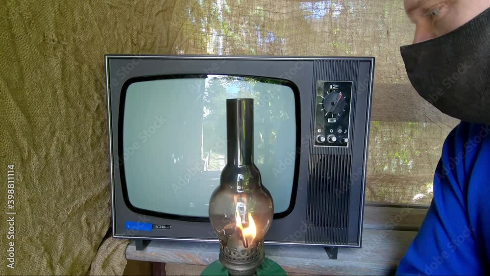 Man in mask against virus and kerosene lamp in his hand turns on an old TV. There is interference on TV, man switches channels and turns off the retro device