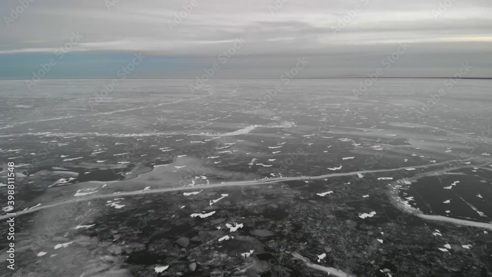 Aerial from drone moving slowly forward over a vast frozen lake covered in black ice and scattered snow drifts. The cracks in the ice create abstract patterns.
Aerial from drone moving slowly forward over a vast frozen lake covered in black ice and scattered snow drifts. The cracks in the ice create abstract patterns.