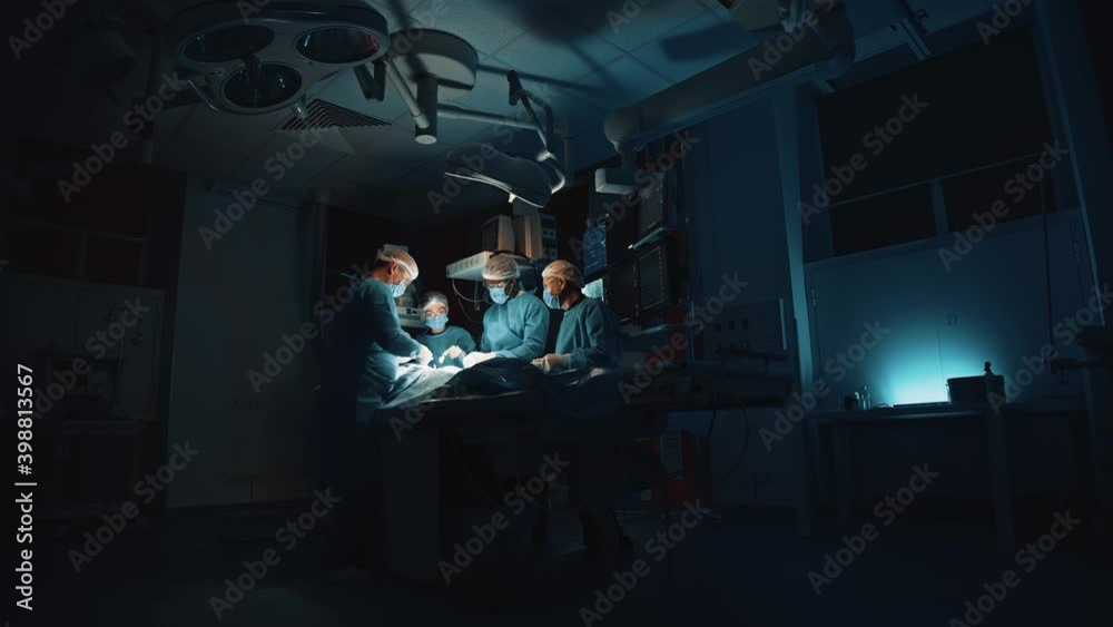 View from the hall of professional doctors at blue medical clothes in the operation theatre under the bright light of a surgical lamp doing urgent operation for the patient at the serious condition View from the hall of professional doctors at blue medical clothes in the operation theatre under the bright light of a surgical lamp doing urgent operation for the patient at the serious condition