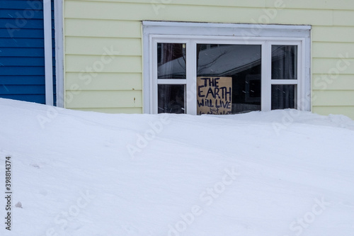 A small cardboard handwritten sign with a message about the earth and the environment in a small window of a yellow building.  Part of the message is covered by a large white snowbank in front.  © Dolores  Harvey