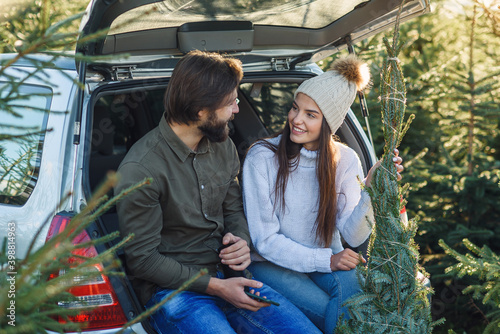 Pleasant couple sitting in car trunk and looking at wonderful christmas tree they choose for holidays at plantings area.