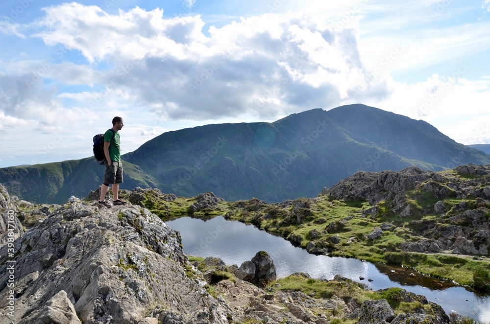 Young hiker at the top of mountain standing next to a little lake and admiring beautiful view. Lake district national park, UK.