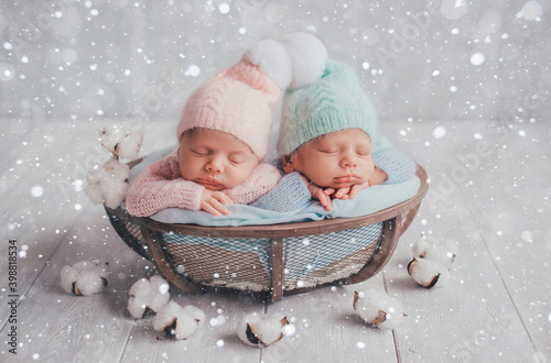 Twins are newborn brother and sister. Newborn girl and boy. Hats with white fur balls sleep sweetly in a basket. Artificial snow.