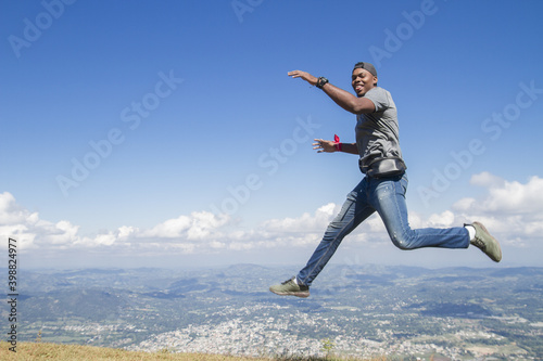 full body shot of one black skinned latin or african american young man outdoors jumping high looking to camera in a summer day vacation trip with nature blue sky in background