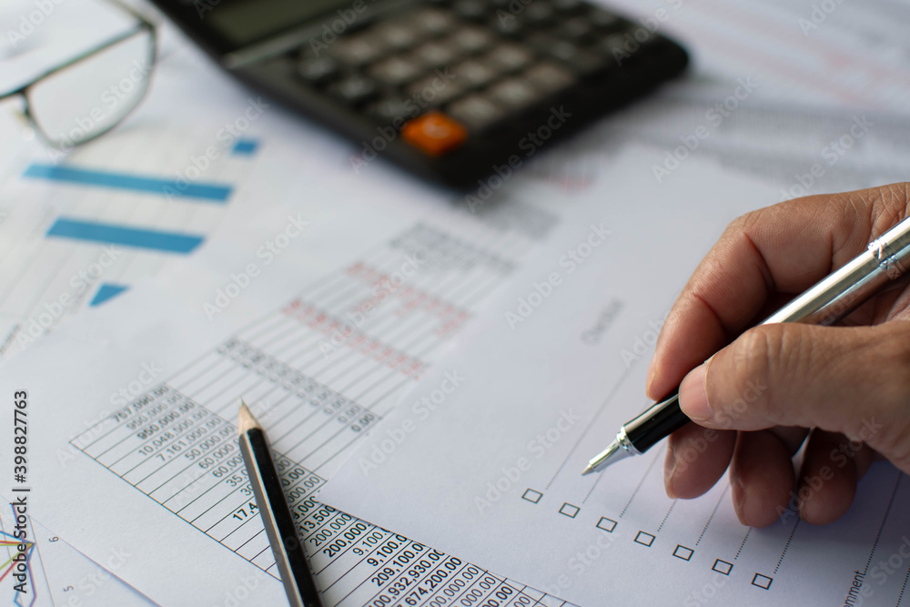 Financial accounting, Closeup, A man holding a pen and checking the list financial transaction, A calculator, pencil, glasses on paper, Graphs showing statistics of financial, Blurred background Financial accounting, Closeup, A man holding a pen and checking the list financial transaction, A calculator, pencil, glasses on paper, Graphs showing statistics of financial, Blurred background