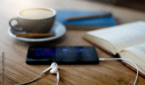 Close-up of white headphones with mobile and book on wooden table. on background blurred coffee drink and blue diary