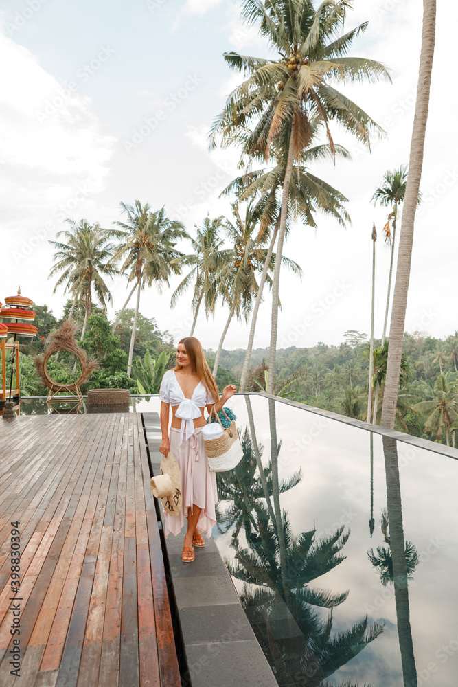 Happy young woman in summer clothes and sun hat with bag walk near swimming pool at beach resort, surrounded with palm trees Happy young woman in summer clothes and sun hat with bag walk near swimming pool at beach resort, surrounded with palm trees